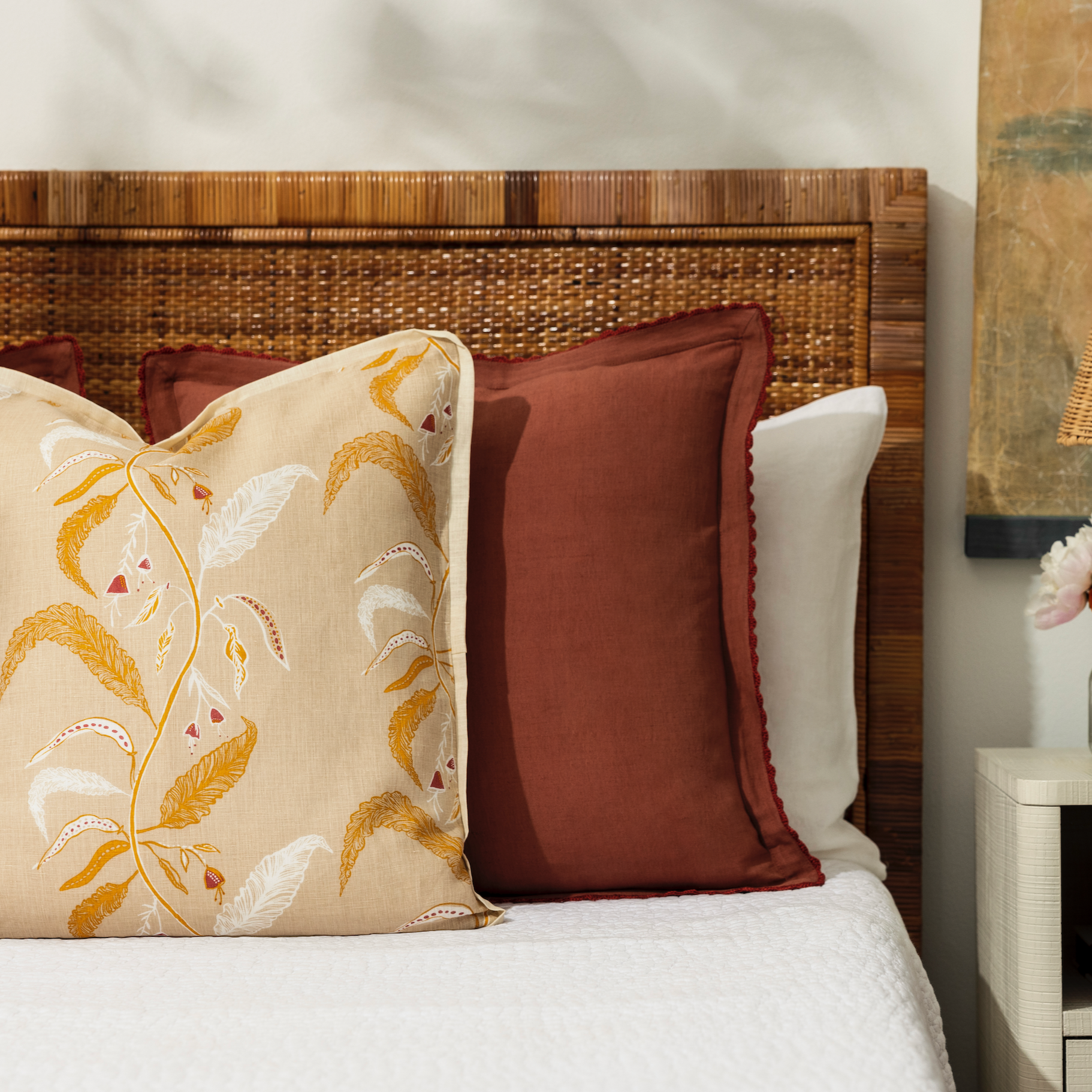 maison venu teracotta pillow propped up on a bed with floral pillow and nightstand with bud vase filled with flowers