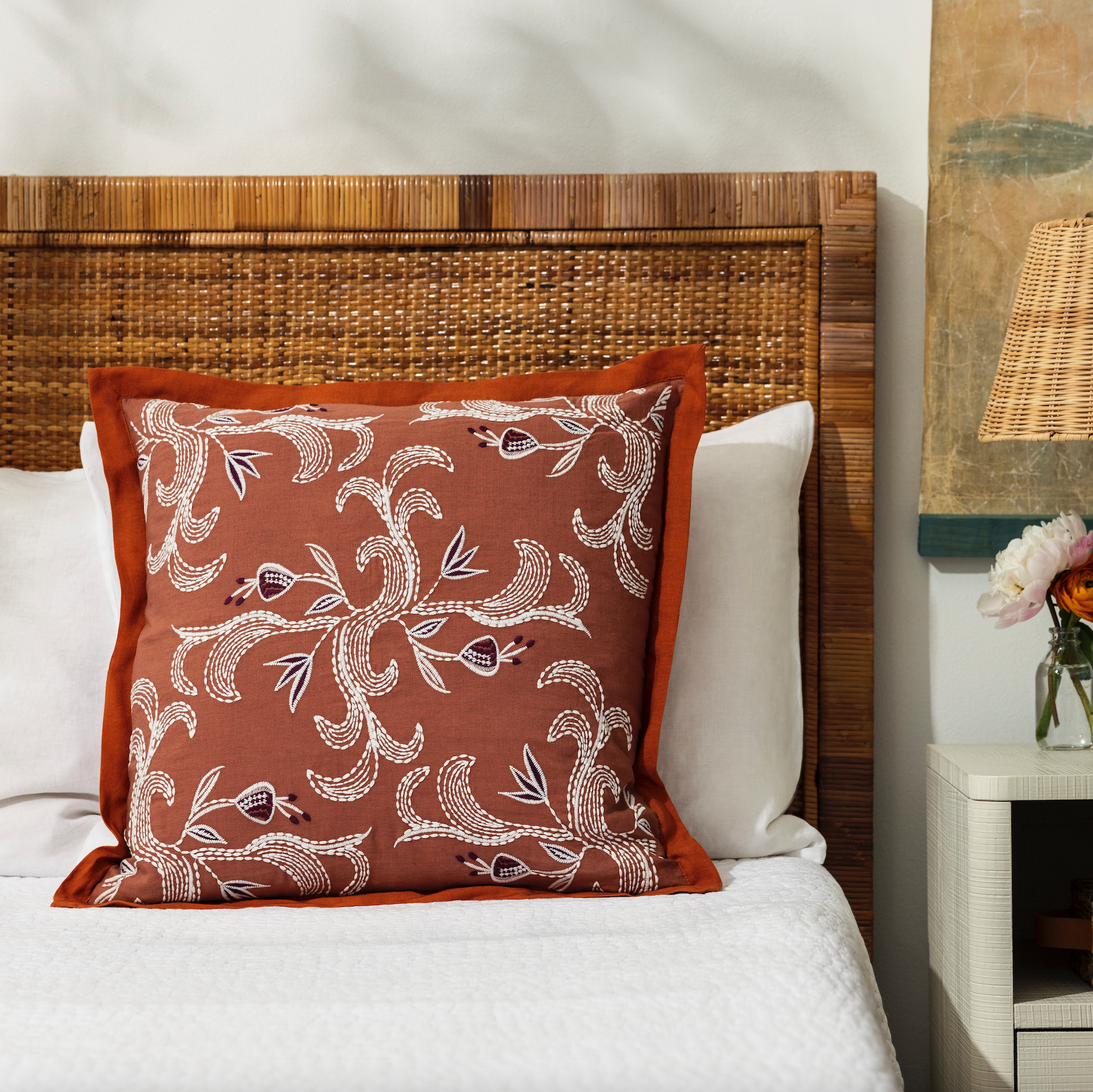 marseille persimmon pillow styled on a bed with white bed linen and a nightstand with flowers in a bud vases and nightstand lamp
