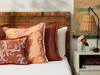 tuileries persimmon pillow stacked on a bed with white bed linen. nightstand with bud vase and flowers and lamp with cane lampshade